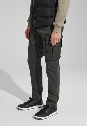 Green cargo pants with multiple pockets and zippers, paired with black sneakers. The outfit features a padded black vest and a light shirt.