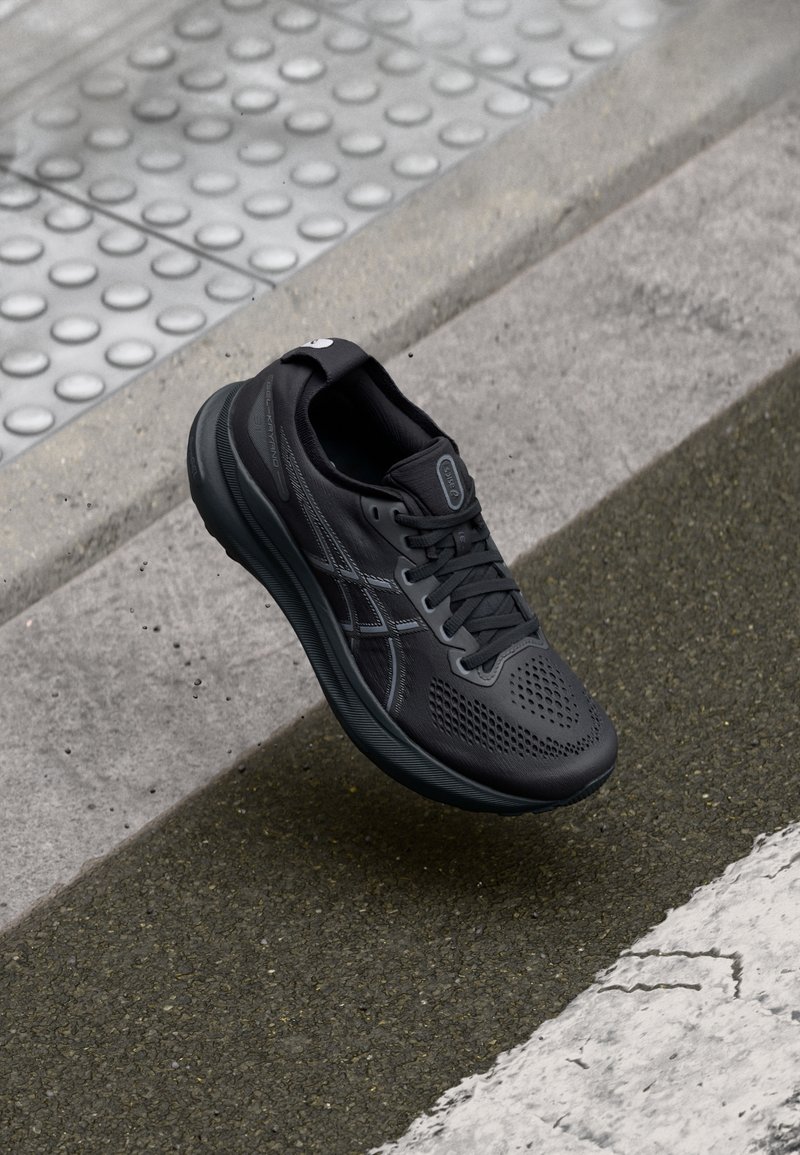 Chaussure de course noire suspendue au-dessus d'un trottoir texturé et d'un bitume, mettant en valeur un design élégant et des caractéristiques de performance.