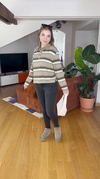 Polo à rayures, coupé, en tons beige et marron, associé à un jean noir et des bottes grises. Tenant un petit sac blanc, à l'intérieur.