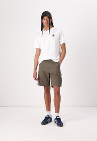 Hvit T-skjorte med kort erme og liten grafikk, olivengrønne cargo-shorts med lommer, svart caps, blå joggesko og hvite sokker.