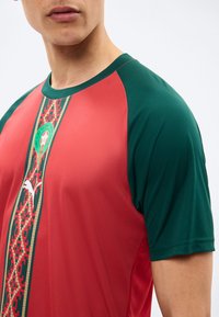 Άνδρας που φοράει κόκκινη και σκούρα πράσινη αθλητική φανέλα με κάθετη ριγέ σχεδίαση και λογότυπο Puma στο μπροστινό μέρος, περικομμένη στη περιοχή του λαιμού και των ώμων.