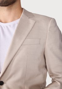 Blazer beige en tissu texturé, avec revers crantés et une poche poitrine. Porté sur un T-shirt blanc. Détail de bouton noir visible.