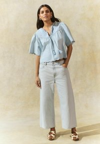 Light blue cropped blouse with wide sleeves, lace detailing, and a tie at the neck. Paired with light blue wide-leg jeans and patterned shoes.