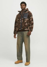 Brown fuzzy jacket with a black chest pocket, paired with light green jeans and tan boots, showcasing a relaxed fit and textured fabric.
