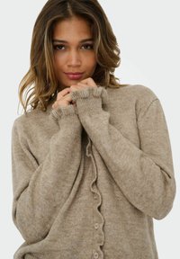 Kafijas krāsas adīta jaka ar tekstūrētu virsmu, ar pogu aizdari un viļņotām malām uz piedurknēm. Mīksts, silts materiāls izceļ sarežģītas šuvju detaļas.