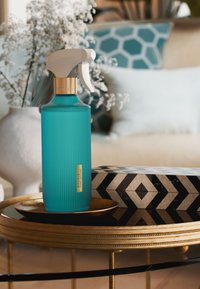 Botella pulverizadora turquesa con diseño acanalado, gatillo blanco y detalle en dorado. Colocada sobre una bandeja dorada junto a una caja con patrón en blanco y negro.