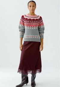 Pull en maille gris avec un motif fair isle rouge et rose, associé à une jupe satin bourgogne avec des bordures en dentelle, et des bottes noires arrivant au genou.