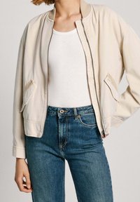 Beżowa kurtka bomberka z prążkowanym kołnierzem, na zamek błyskawiczny i bocznymi kieszeniami, w zestawie z niebieskimi dżinsami. Gładka tekstura materiału i swobodny design.