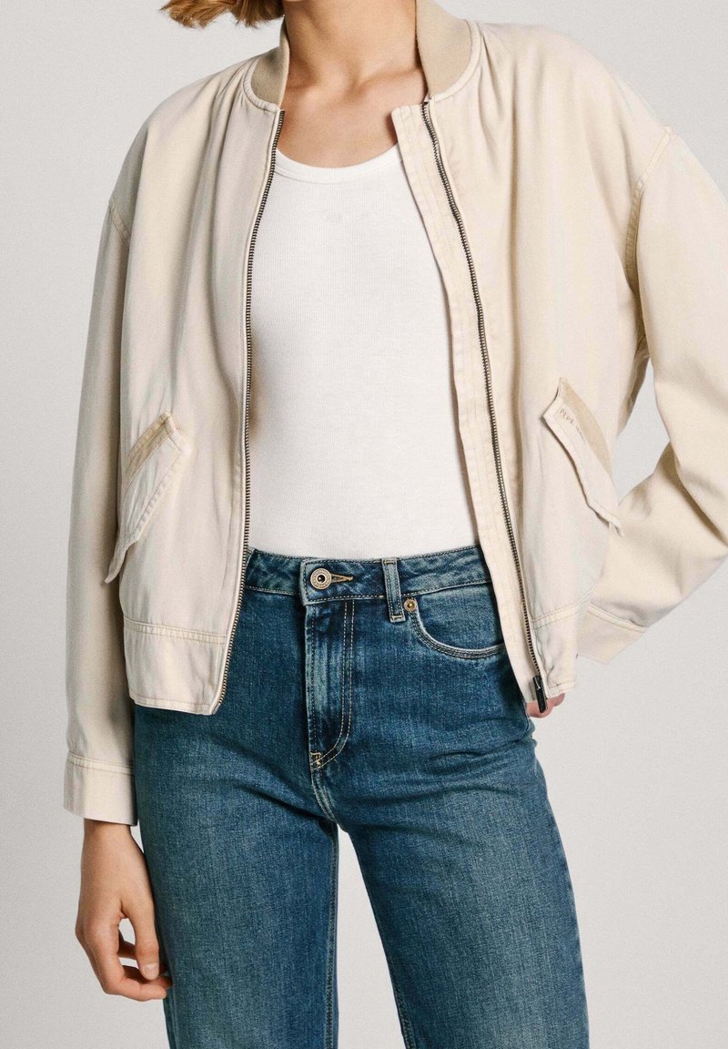Beżowa kurtka bomberka z prążkowanym kołnierzem, na zamek błyskawiczny i bocznymi kieszeniami, w zestawie z niebieskimi dżinsami. Gładka tekstura materiału i swobodny design.