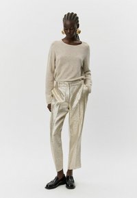 Šviesiai rudas ilgos rankovės megztinis derinamas su blizgiais auksinės tekstūros kelnėmis. Juodi loafers bateliai užbaigia įvaizdį, akcentuodami kontrastingas medžiagas.