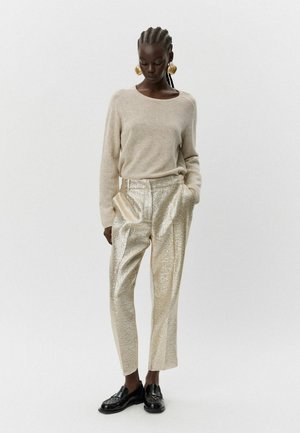 Šviesiai rudas ilgos rankovės megztinis derinamas su blizgiais auksinės tekstūros kelnėmis. Juodi loafers bateliai užbaigia įvaizdį, akcentuodami kontrastingas medžiagas.