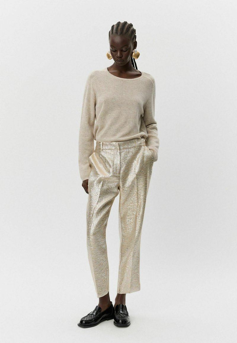 Šviesiai rudas ilgos rankovės megztinis derinamas su blizgiais auksinės tekstūros kelnėmis. Juodi loafers bateliai užbaigia įvaizdį, akcentuodami kontrastingas medžiagas.