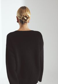 Schwarzer Pullover aus weichem, strukturiertem Stoff mit legerem Sitz und überschnittenen Schultern. Saum und Bündchen sind gerippt. Das Model wird von hinten betrachtet.