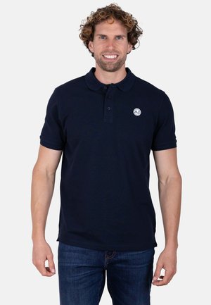Hombre sonriente con el pelo rizado, vestido con un polo azul marino oscuro con un pequeño logotipo blanco y vaqueros azules, de pie frente a un fondo claro y liso.