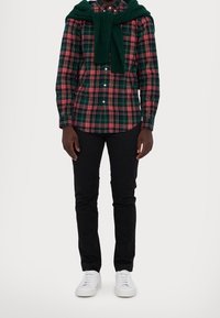 Camicia a scacchi verde, rossa e bianca, con un maglione verde appoggiato sulle spalle. Pantaloni neri e sneaker bianche completano l'outfit.