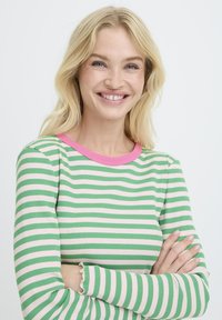 Lächelnde blonde Frau mit gekreuzten Armen, die ein langärmliges Oberteil mit grün-weißen Streifen und einem pinken Ausschnitt trägt, vor einem weißen Hintergrund.