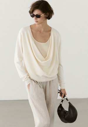 Frau mit kurzen Haaren, die dunkle Sonnenbrille trägt, cremefarbenes, locker fallendes Oberteil und hellbeige Hose, hält eine schwarze Handtasche vor schlichtem Hintergrund.