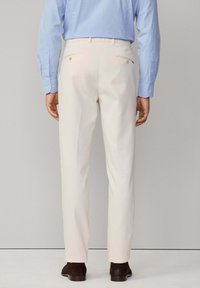 Pantaloni color crema con gamba dritta, presentano due tasche posteriori con bottoni e una finitura del tessuto liscia, indossati con una camicia azzurro chiaro.