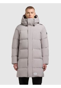 Gai pelēks pūkains mētelis ar augstu apkakli un lielu kapuci. Iezīmējas ar elegantu dizainu, horizontālām quiltētām sekcijām un sānu kabatām.