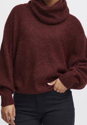 Person trägt einen bordeauxfarbenen, flauschigen Rollkragenpullover mit langen Ärmeln und schwarze Hose, Hand ruht in der Tasche.