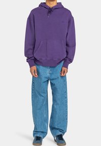 Persona de pie vestida con una sudadera morada, vaqueros claros holgados y zapatillas azules frente a un fondo claro y uniforme.