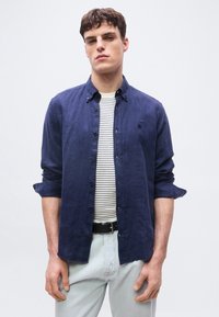 Νέος άνδρας φοράει μπλε πουκάμισο με κουμπιά πάνω από λευκό και μπλε ριγέ μπλουζάκι, συνδυασμένο με ανοιχτόχρωμα τζιν και μαύρη ζώνη.