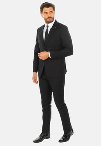 Completo da uomo nero con blazer strutturato e pantaloni slim-fit. Scarpe nere lucide e una camicia bianca completano il look.