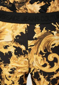 Μαύρο και χρυσό παντελόνι με μπαρόκ σχέδιο και μαύρη ζώνη μέσης με ανάγλυφο κείμενο «Versace Jeans Couture».