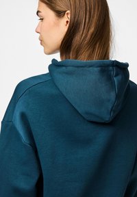 Huvtröja i en mörk teal färg med en mjuk textur. Har en känguruficka och avslappnad passform, visar bakvyn.
