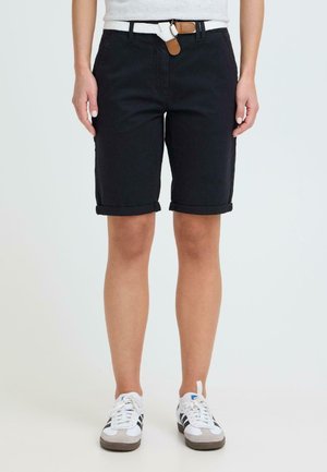 Person iført svarte shorts med oppbrettet kant, hvitt flettet belte og hvite joggesko med svarte striper, stående mot en ensfarget bakgrunn.