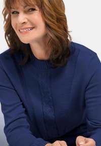 Vrouw met schouderlang bruin haar glimlacht, gekleed in een blauwe gebreide trui met een gestructureerd verticaal patroon aan de voorkant.
