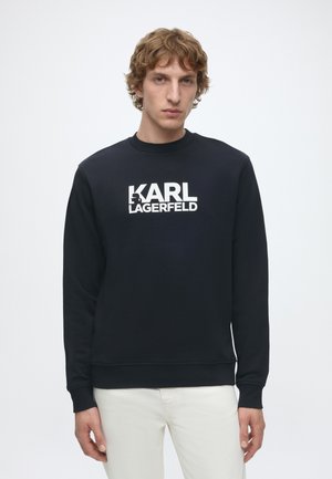 Młody mężczyzna z kręconymi brązowymi włosami, ubrany w czarną bluzę z logo "KARL LAGERFELD" i białe spodnie, stoi na jednolitym tle.