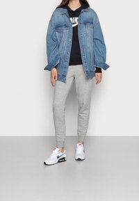 Osoba oblečená v přerostlé modré džínové bundě, černé mikině Nike, šedých teplácích a bílých teniskách Nike Air stojí na šedé podlaze.