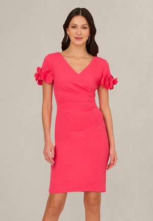 Žena stojící před jednobarevným pozadím, oblečená v růžových šatech délky ke kolenům s výstřihem do V a krátkými rukávy zdobenými velkými květinovými aplikacemi.