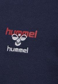 Navyblauwe stof heeft prominente geborduurde tekst "hummel" in rood en wit, samen met een gestileerd bijlogo in het wit.