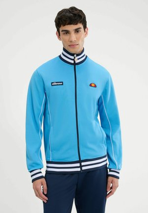 Junger Mann trägt eine hellblaue Jacke mit Reißverschluss, weiß-navy gestreiften Kragen, Bündchen und Saum, mit ellesse-Logo-Patches auf der Brust.