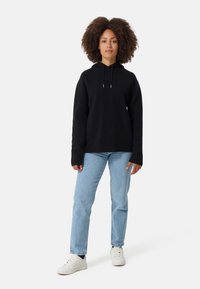 Felpa nera con cappuccio realizzata in un tessuto testurizzato, caratterizzata da una vestibilità ampia e maniche lunghe, abbinata a jeans azzurri chiari e sneakers bianche.