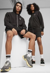 Sorte zip-up hættetrøjer og shorts, med grå og sorte sneakers. Modellerne er placeret på en pedestal, hvilket fremhæver detaljerne i tøjet og skoene.