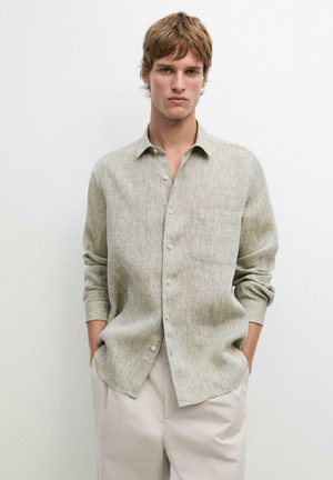 Young man with light brown hair wearing a beige linen button-up shirt and light-colored pants, standing with hands in pockets against plain background.