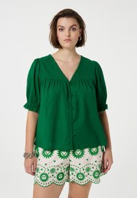 Groene linnen blouse met korte poff mouwen en een V-hals, voorzien van decoratieve knopen en bies. Gecombineerd met gestreepte crèmekleurige shorts.
