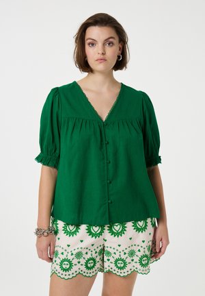 Grüne Leinenbluse mit kurzen Puffärmeln und V-Ausschnitt, ausgestattet mit dekorativen Knöpfen und Besatz. Kombiniert mit gemusterten cremfarbenen Shorts.