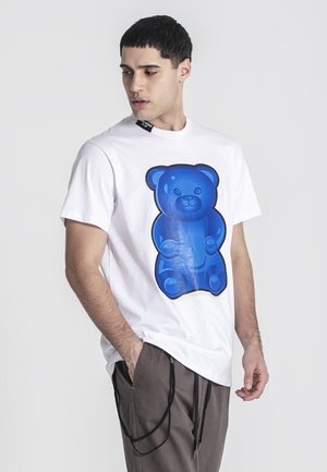 Jeune homme aux cheveux courts et foncés, portant un t-shirt blanc avec un grand motif ours en gélatine bleu, et un pantalon marron avec des cordons noirs.