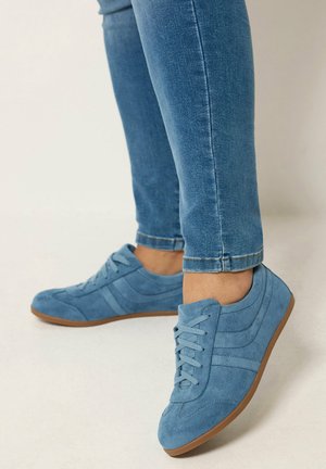 Personne portant des baskets en daim bleu clair avec des semelles marron et un jean bleu, debout sur un fond neutre clair.