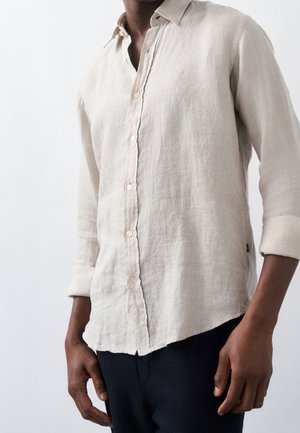 Homme portant une chemise en lin beige à manches longues avec les manches retroussées et un pantalon sombre sur un fond clair uni.