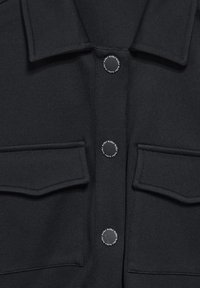 Sort jakke med struktureret krave, tre trykknapper ned langs midten, og to forsæde lommer.