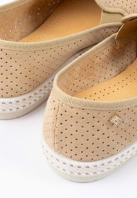 Beige geperforeerde instappers met een zachte suède bovenkant, voorzien van een witte geweven tussenzool en een klein gouden accent op de hiel.