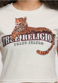 Kremasta majica s kratkim rukavima koja ima grafiku leoparda izrađenu od rhinestona, s natpisom "TRUE RELIGION BRAND JEANS" u podebljanom fontu.