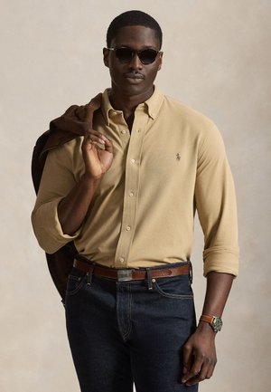 Mand iført beige skjorte med knapper, mørke jeans, solbriller og ur, der holder en brun jakke over skulderen mod en ensfarvet baggrund.
