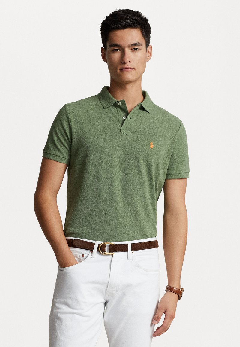 Polo shirt de algodón verde con collar, con un pequeño logo naranja en el pecho. Combinado con pantalones blancos y un cinturón marrón.
