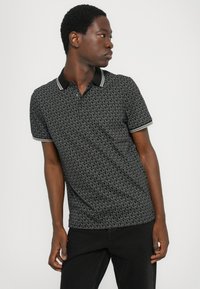 Polo noir avec un motif monogramme MK sur l'ensemble, col contrasté avec deux boutons, et manches courtes avec des accents rayés.